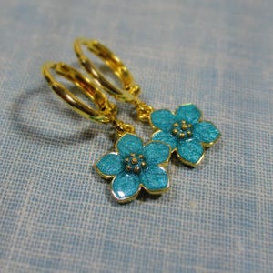 Auf einem hellblauen Untergrund liegt ein Paar kleine goldene Creolen. An jeder Creole hängt eine blaue Blüte.