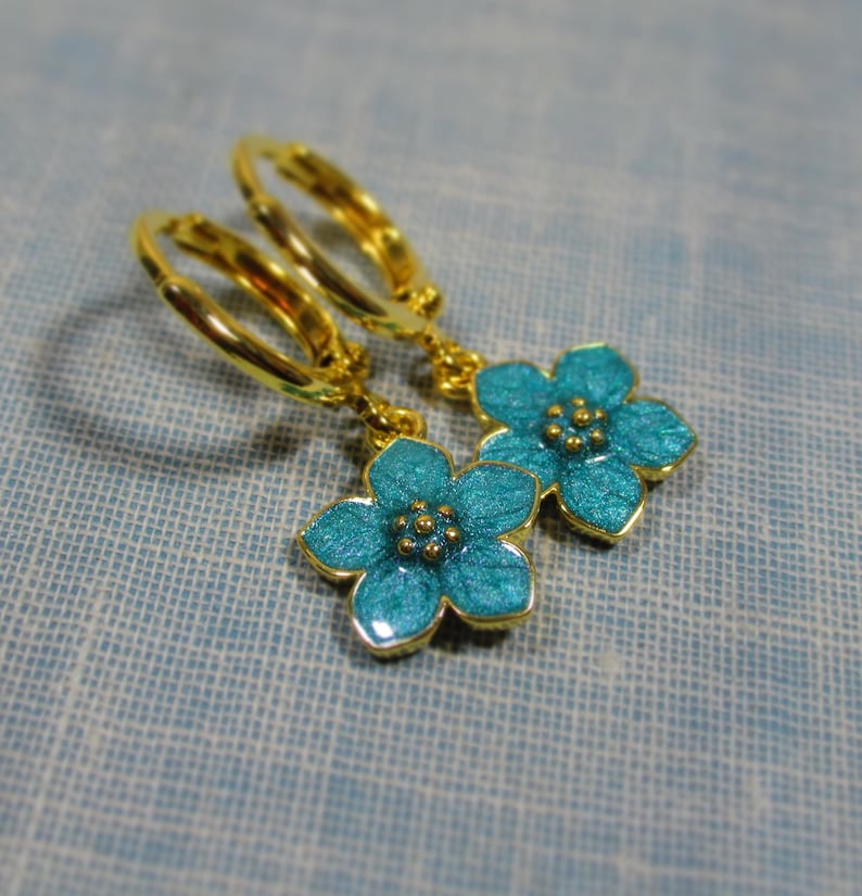 Auf einem hellblauen Untergrund liegt ein Paar kleine goldene Creolen. An jeder Creole hängt eine blaue Blüte.