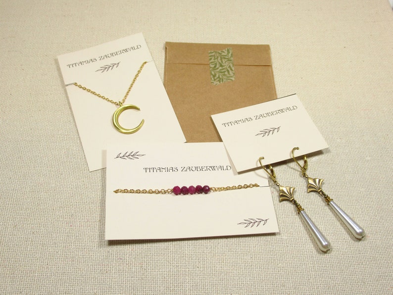 Auf einem beigenen Untergrund liegen Beispiele für die Verpackung der Schmuckstücke von Titanias Zauberwald: eine Kette mit Mondanhänger an einer hellbeigenen Karte, ein Armband mit dunkelroten Perlen auf einer hellbeigenen Karte, ein paar Ohrringe mit langen weißen Tropfenperlen an einer hellbegeienen Karte und ein braunes Papiertütchen.