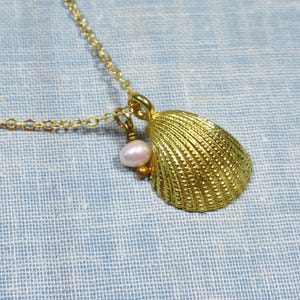 May include: A gold-colored seashell pendant necklace with a small, light pink pearl charm. The seashell has a textured surface and is suspended from a delicate gold chain. The necklace is displayed on a light blue background.