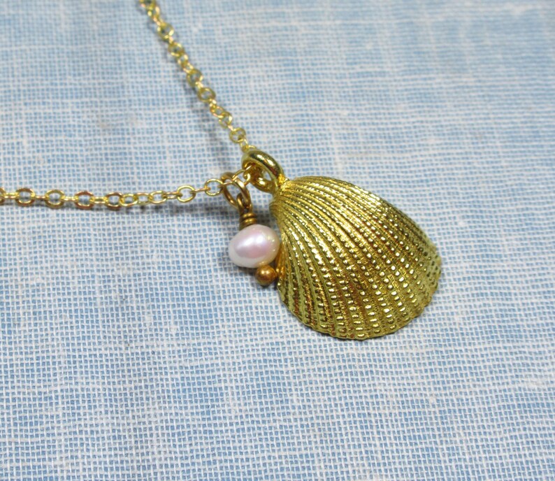 May include: A gold-colored seashell pendant necklace with a small, light pink pearl charm. The seashell has a textured surface and is suspended from a delicate gold chain. The necklace is displayed on a light blue background.