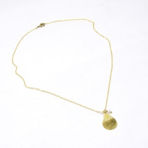 May include: A gold-colored necklace featuring a seashell pendant and a delicate chain. The pendant has a textured, fan-like design, and a small pearl is attached. The necklace is displayed against a white background.