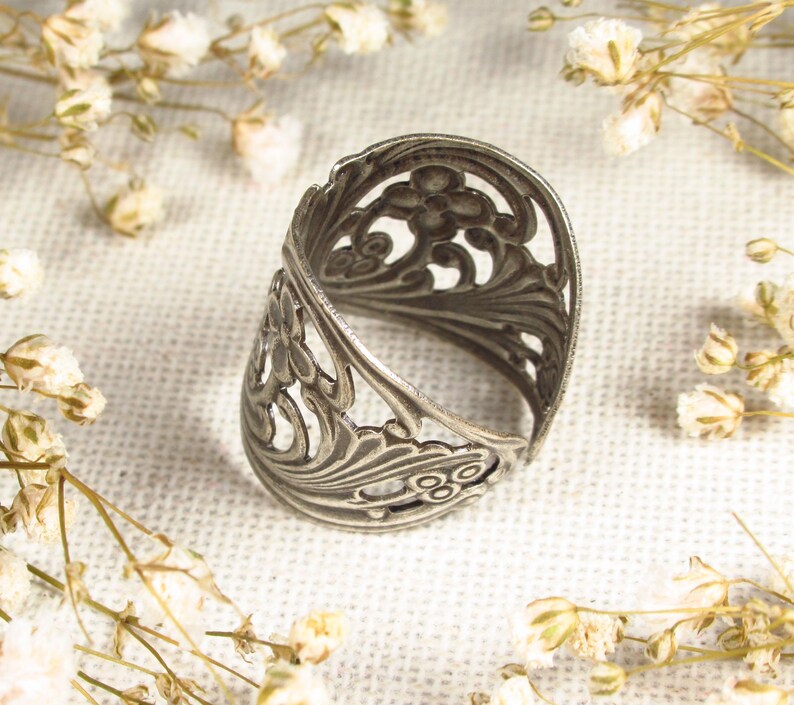 May include: A silver filigree ring with a delicate floral design. The ring is open and has a wide band.
