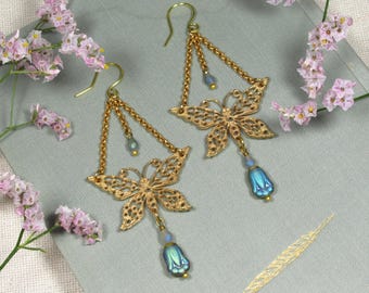 Puede incluir: Un par de pendientes de mariposa dorados con detalles de lágrima azul iridiscente. Los pendientes presentan alas de mariposa delicadas y cuelgan de una cadena de tres hebras.
