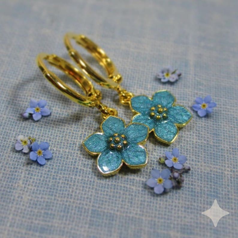 Auf einem hellblauen Untergrund liegt ein Paar kleine goldene Creolen. An jeder Creole hängt eine blaue Blüte. Um die Ohrringe herum liegen kleine echte Vergissmeinnicht-Blüten.