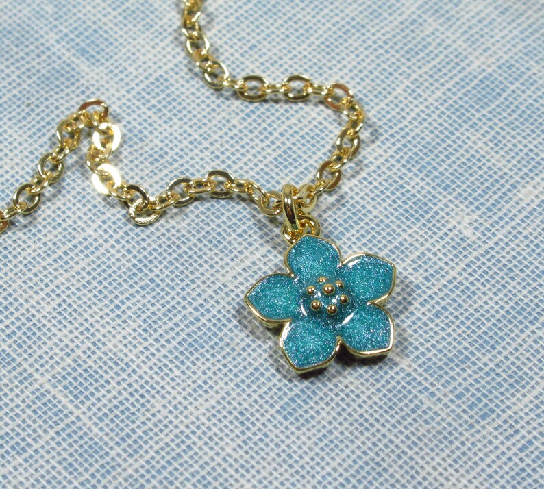 Auf einem hellblauen Hintergrund liegt eine goldene Halskette, von der ein kleiner Ausschnitt zu sehen ist. An der Halskette befindet sich ein Anhänger in Form einer hellblauen Blüte.