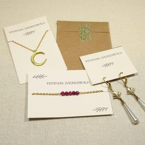 Auf einem beigenen Untergrund liegen Beispiele für die Verpackung der Schmuckstücke von Titanias Zauberwald: eine Kette mit Mondanhänger an einer hellbeigenen Karte, ein Armband mit dunkelroten Perlen auf einer hellbeigenen Karte, ein paar Ohrringe mit langen weißen Tropfenperlen an einer hellbegeienen Karte und ein braunes Papiertütchen.
