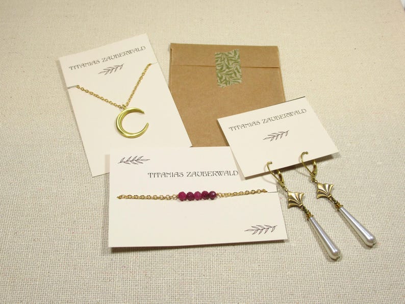 Auf einem beigenen Untergrund liegen Beispiele für die Verpackung der Schmuckstücke von Titanias Zauberwald: eine Kette mit Mondanhänger an einer hellbeigenen Karte, ein Armband mit dunkelroten Perlen auf einer hellbeigenen Karte, ein paar Ohrringe mit langen weißen Tropfenperlen an einer hellbegeienen Karte und ein braunes Papiertütchen.