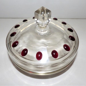 May include: A clear glass candy dish with a lid. The round dish has a faceted design and is decorated with small, oval, ruby-red accents around the rim. The lid has a clear, faceted knob.