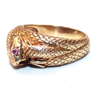 May include: A gold ring shaped like a coiled snake with textured scales covering the band. The snake head features a small, round, vibrant pink gemstone set as an eye, with the body wrapping around to form the circular band of the ring.