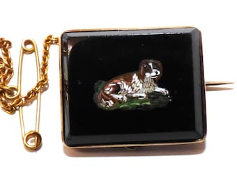 May include: A gold brooch with a black square cameo featuring a mosaic of a brown and white dog. The brooch has a safety pin and chain.