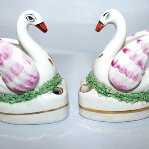 Puede incluir: Par de figuras de cisnes de cerámica. Los cisnes son blancos con detalles de alas rosas y moradas y picos rojos. Se posan sobre una base blanca con una banda dorada y adornos verdes. Decoración del hogar.