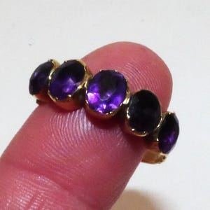 Puede incluir: Un anillo de oro con cinco piedras preciosas ovaladas de color púrpura. Las piedras están engastadas en una banda de oro, creando una fila continua de color. El anillo se muestra en un dedo, mostrando su diseño y color.
