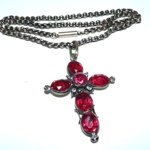 May include: A silver necklace featuring a cross pendant with red gemstones. The cross design incorporates oval-shaped red stones and smaller clear stones. The chain is made of interconnected links.