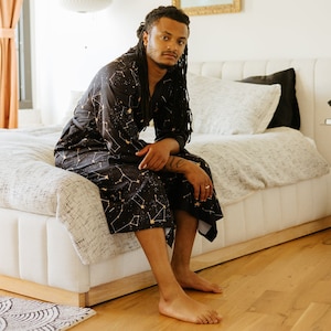 Könnte beinhalten: Eine Person in einer schwarzen und goldenen Robe mit einem Sternenbildmuster sitzt auf einem weißen Bett mit einer weißen Decke. Die Person hat lange Dreadlocks und schaut in die Kamera.