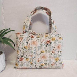 May include: A quilted tote bag featuring a cream-colored fabric base with a repeating floral pattern in shades of yellow, pink, orange, and green. The bag has two matching fabric handles and vertical stitching lines across the entire surface.