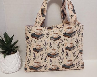 Puede incluir: Un bolso de tela acolchada con un patrón repetitivo de tazas de té humeantes sobre pilas de libros, intercaladas con ramitas de lavanda sobre un fondo crema. El bolso tiene dos asas de tela a juego y una forma rectangular.