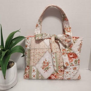 May include: A quilted tote bag with a patchwork design featuring floral patterns in shades of pink, peach, and green on a white background. The bag has matching handles and a decorative bow. The bag is approximately 10 inches wide and 8 inches tall.