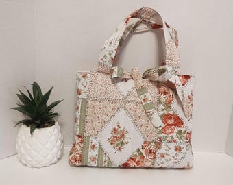 Puede incluir: Un bolso de tela acolchada con un diseño de patchwork de motivos florales en tonos rojo, verde y crema. El bolso incluye dos asas superiores y un lazo de tela decorativo atado en la parte delantera, con costuras visibles en la superficie.