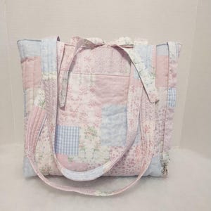 May include: A quilted tote bag featuring a patchwork design in soft pastel shades of pink, light blue, and white. The fabric displays various floral patterns and a small gingham check print. It includes two fabric shoulder straps and a metal clip.