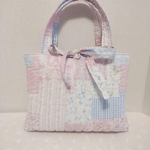 May include: A quilted tote bag featuring a patchwork design of various floral, striped, and gingham fabrics in soft shades of pink, light blue, and white. The bag includes two matching fabric handles and a decorative fabric tie at the center top.