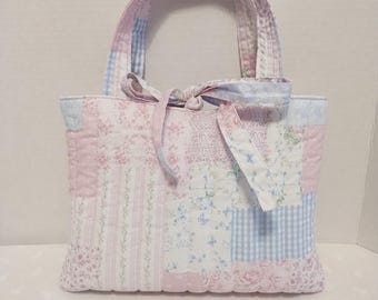 Puede incluir: Un bolso de mano acolchado con un diseño de patchwork en telas de color rosa claro, azul y blanco. El patrón incluye estampados florales, rayas y cuadros vichy. El bolso tiene dos asas de tela y un cierre de lazo decorativo en el centro.