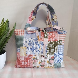 May include: A quilted tote bag featuring a patchwork design of various floral, gingham, and striped patterns in shades of blue, pink, green, and white. The bag includes two matching fabric handles and a decorative fabric tie at the centre top.