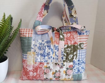 Puede incluir: Un bolso tote acolchado con un diseño de patchwork que combina varios estampados florales, de cuadros vichy y de rayas en tonos azul, rosa, verde y blanco. El bolso incluye dos asas de tela a juego y un lazo decorativo en la parte superior.
