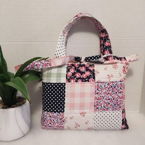 May include: A patchwork tote bag with a variety of floral, polka dot, and gingham patterns in pink, navy, and white. The bag has a square shape with a matching handle and is likely handmade.