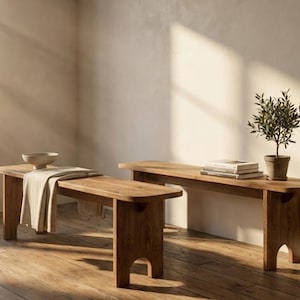 May include: Two wooden benches with a natural finish are displayed in a sunlit room. The benches have a simple, minimalist design. One bench has a bowl and a folded cloth. The other bench has a small potted plant and books.