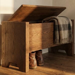 Peut inclure: Un banc de rangement en bois avec un couvercle à charnière, ouvert pour révéler une couverture pliée. Le banc est en bois brun clair et de forme rectangulaire. Une paire de bottes en cuir marron est visible sous le banc.