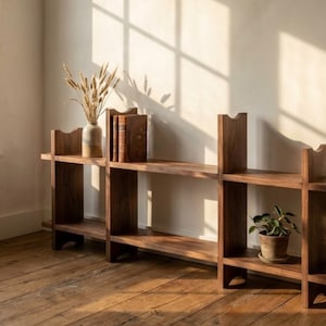 May include: A wooden bookshelf with two shelves and a unique cutout design. The shelves hold a vase with dried plants, several books, and a potted plant. The bookshelf is made of dark wood and sits on a wooden floor.