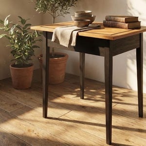 May include: A small, square wooden table with a dark brown frame and a light brown tabletop. The table has four legs and is topped with a stack of books, bowls, and a folded cloth. Potted plants are in the background.