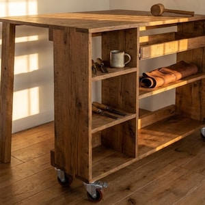 May include: A wooden workbench with a tabletop and integrated shelving. A mallet rests on top, with tools and a mug on the shelves. The unit is on wheels. The wood has a rustic appearance.