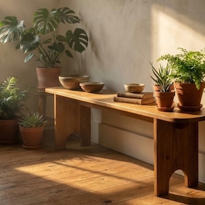 Peut inclure: Un banc en bois avec une finition naturelle, accueillant plusieurs plantes en pot et de petits bols en bois. Les plantes comprennent un grand Monstera, des fougères et des plantes succulentes. Le banc est placé contre un mur neutre.