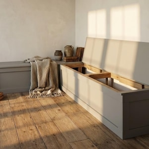 Könnte beinhalten: Eine hellgraue Aufbewahrungsbank mit einem Klappdeckel, der sich öffnet, um das Innere freizulegen. Links befindet sich eine zweite, kleinere Bank. Eine beige Decke und dekorative Töpferwaren sind in der Nähe. Die Bänke stehen auf einem Holzboden.