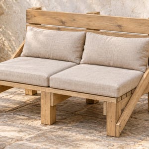 May include: A two-seat bench constructed from light brown wooden planks featuring a slatted backrest and angled side supports. The bench includes two thick, rectangular seat cushions and two matching back cushions in a neutral beige fabric.