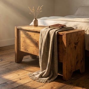 Puede incluir: Un cofre de almacenamiento de madera rústica con una manta encima. Un pequeño jarrón con plantas secas y un libro encuadernado en cuero están encima. El cofre se coloca al pie de una cama con ropa de cama de color neutro.