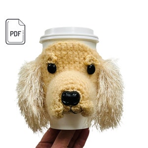 Könnte beinhalten: Häkel-Becherwärmer in Form eines Golden Retriever-Hunde-Kopfes. Der Wärmer hat eine braune Nase, schwarze Augen und flauschige weiße Ohren.