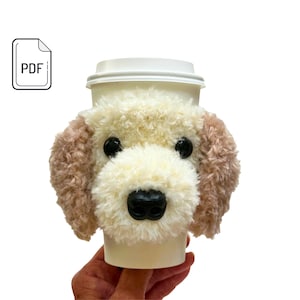 May include: A white coffee cup with a beige and brown dog-shaped cozy. The cozy has a fluffy texture and black eyes and nose.
