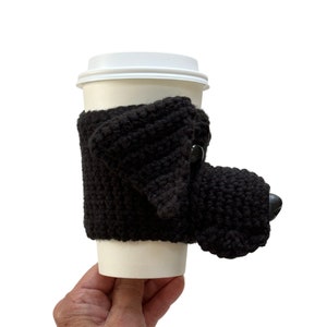May include: A black Lab crocheted coffee cup cozy shaped like a realistic dog head. The cozy has a black plastic dog nose and black ears.