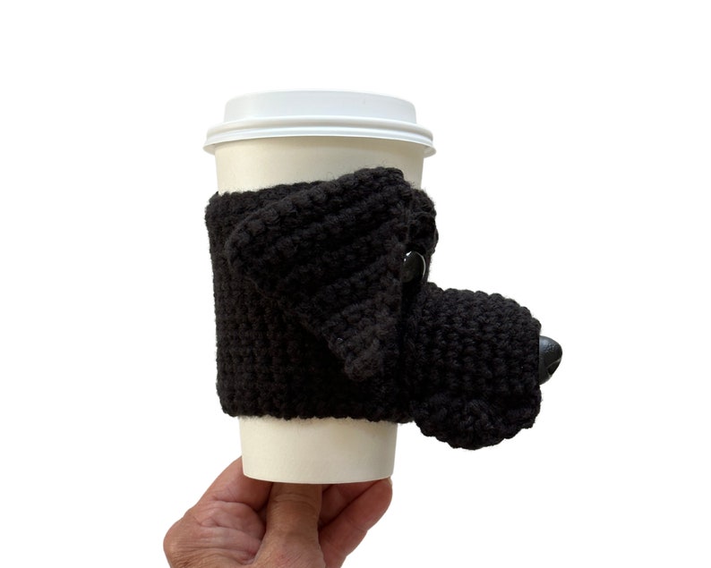 May include: A black Lab crocheted coffee cup cozy shaped like a realistic dog head. The cozy has a black plastic dog nose and black ears.