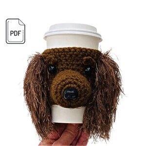 May include: Crocheted brown dog-shaped coffee cup cozy. The cozy has a brown crocheted face with black eyes and nose.
