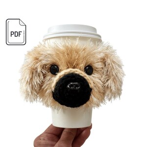 May include: A crocheted dog-shaped coffee cup cozy in beige and black. The cozy is shaped like a dog's head with floppy ears and a black nose.