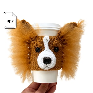 May include: Crocheted coffee cup cozy featuring a brown and white dog with floppy ears. The cozy is made with brown yarn and has a white face with black eyes and a black nose.