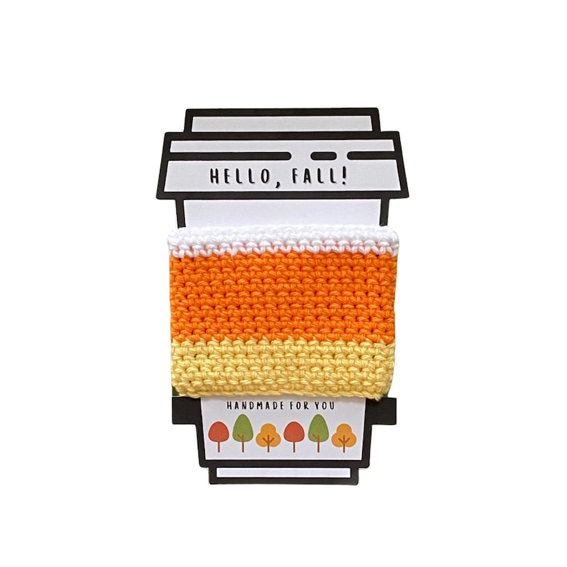 May include: A crocheted coffee cup cozy in orange, yellow, and white with the text "HELLO, FALL!" and "HANDMADE FOR YOU" with a row of stylized trees below.