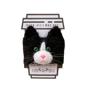 May include: Black and white crocheted cat mug cozy with green eyes and pink nose. The cozy has the text "Purr-fect Mug Hug" and "Handmade Cuteness" on it.