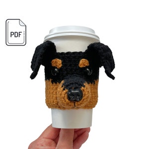 May include: Crocheted coffee cup cozy in the shape of a black and brown dog's face. The cozy has a black nose, brown fur, and black eyes.