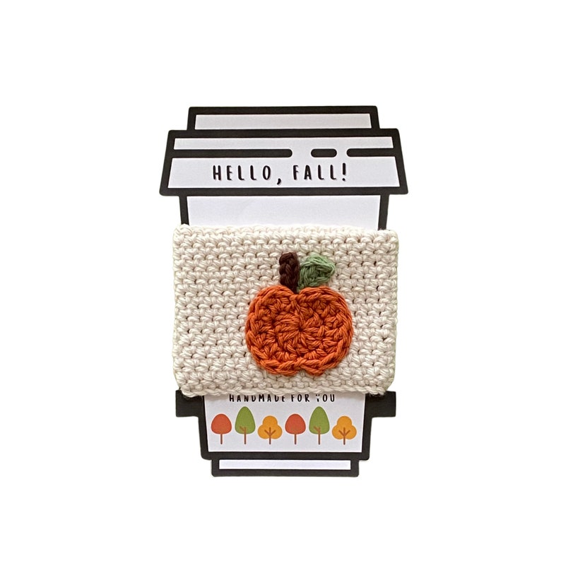 May include: A white crocheted coffee cup cozy with an orange pumpkin design. The cozy has the text "HELLO, FALL!" and "HANDMADE FOR YOU" on it.