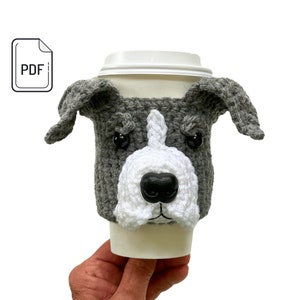 May include: A grey and white crocheted dog-faced coffee cup cosy. The cosy has black button eyes and a black nose. The PDF icon is in the upper left corner.